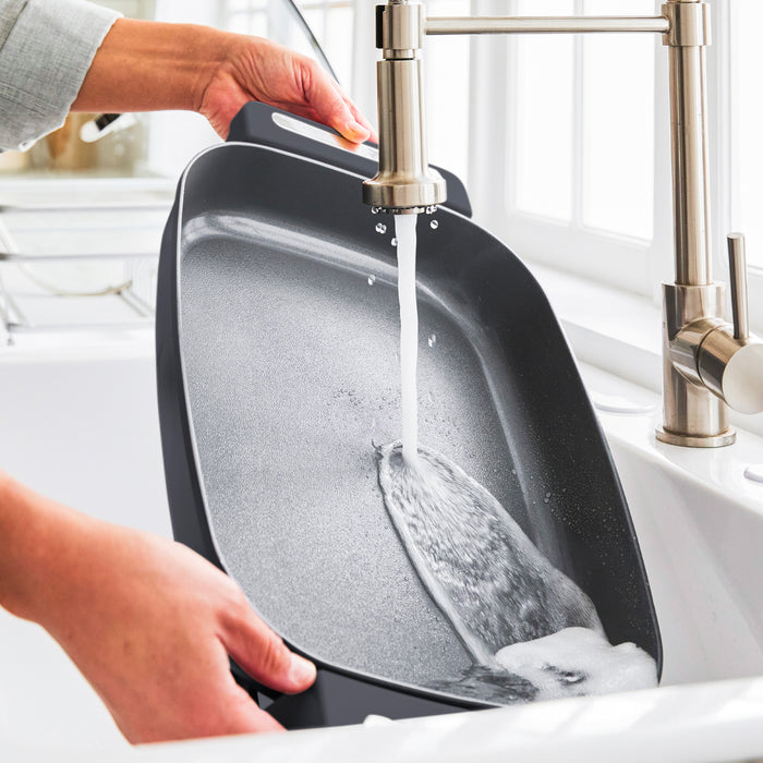 Person washing a graphite 7-quart skillet in a kitchen sink by a window after cooking.
