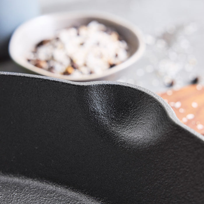 Close-up of the spout on a Bobby Flay by GreenPan 8" and 10" Cast Iron Frypan Set, highlighting its spacious cooking surface, with a blurred bowl of coarse salt and pepper in the background—perfect for any modern kitchen.