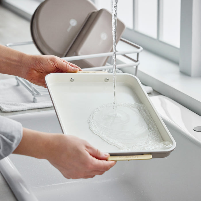 Hands rinsing a latte-colored Reserve Bakeware 13″ x 9″ quarter sheet pan under a kitchen faucet.