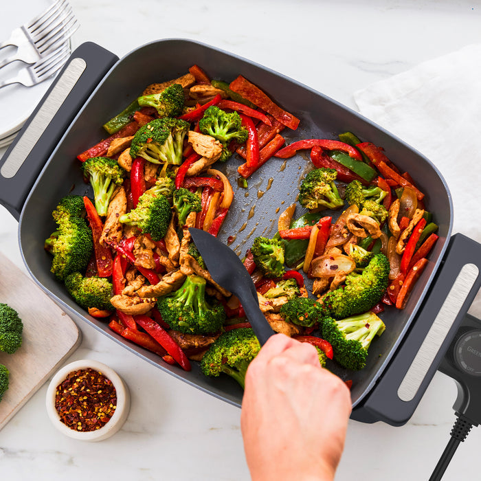 Chicken, broccoli, and peppers being stirred in a graphite 7-quart skillet on a white countertop.