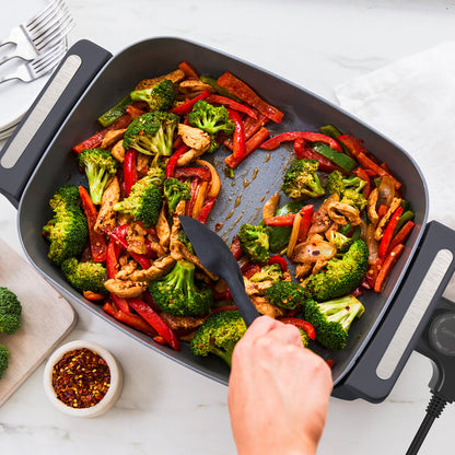 Chicken, broccoli, and peppers being stirred in a graphite 7-quart skillet on a white countertop.