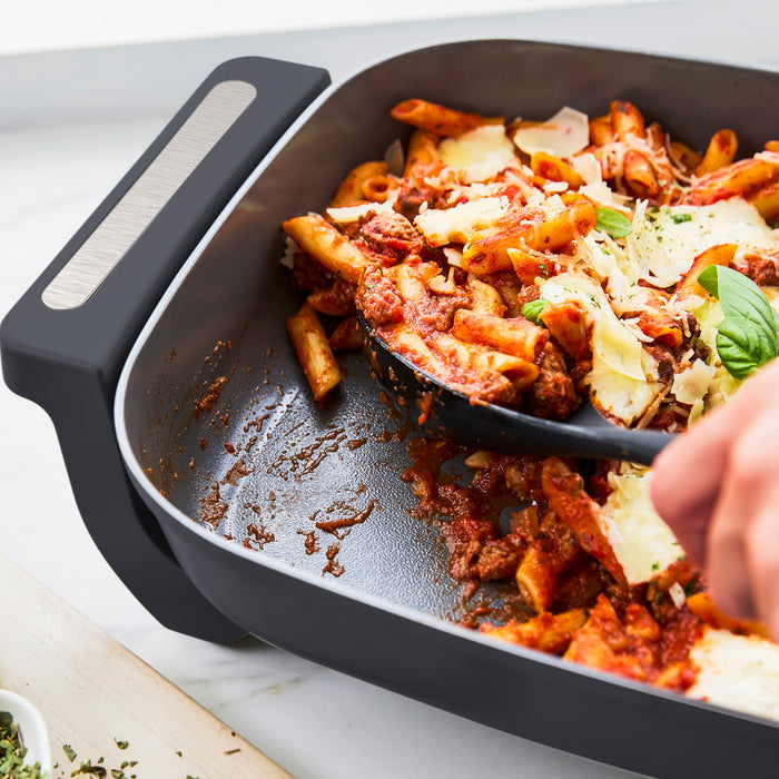 Baked pasta being served from a graphite-colored 7-quart skillet with a spatula. Only the hand and pan are visible.