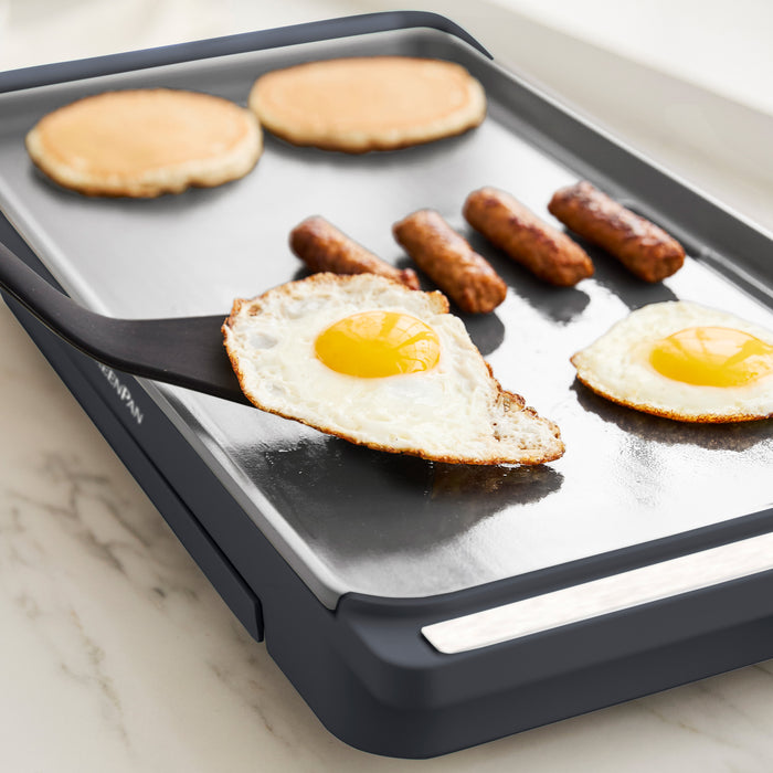 A close-up of the Advanced Griddle with Warming Drawer in graphite on a kitchen counter, cooking eggs, sausages, and pancakes. A spatula lifts an egg, showcasing its family-sized brunch capacity.