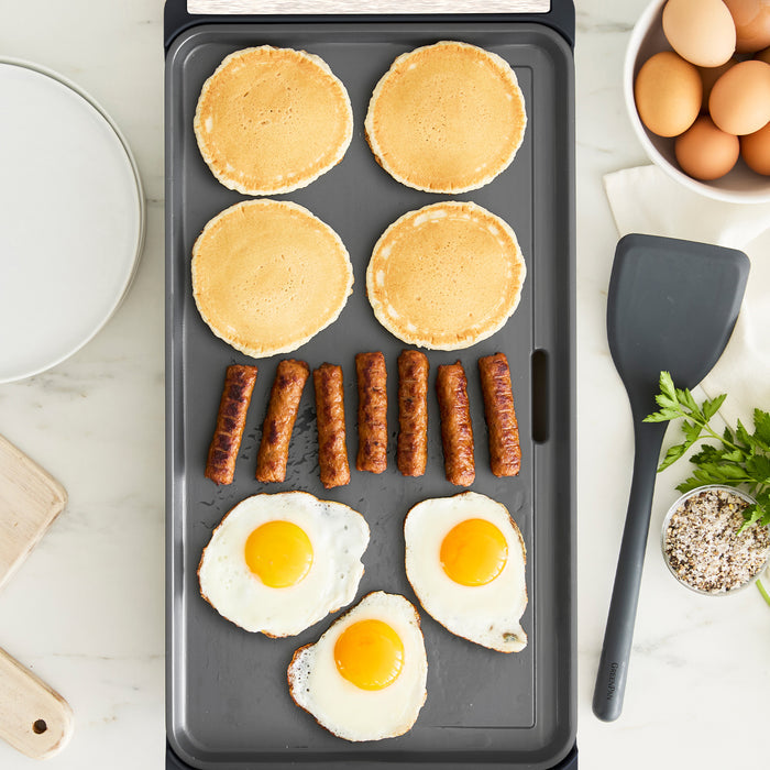 A family brunch cooks on the Advanced Griddle with Warming Drawer | Graphite: four pancakes, six sausage links, and three sunny-side-up eggs sizzle while a plate, bowl of eggs, spatula, herbs, and seasoning rest on the marble countertop.
