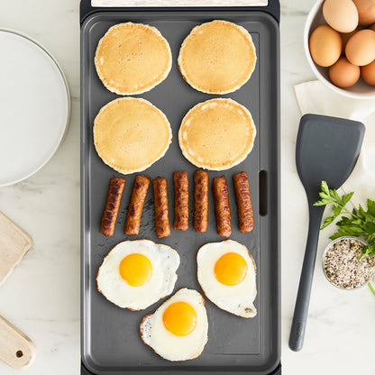 A family brunch cooks on the Advanced Griddle with Warming Drawer | Graphite: four pancakes, six sausage links, and three sunny-side-up eggs sizzle while a plate, bowl of eggs, spatula, herbs, and seasoning rest on the marble countertop.