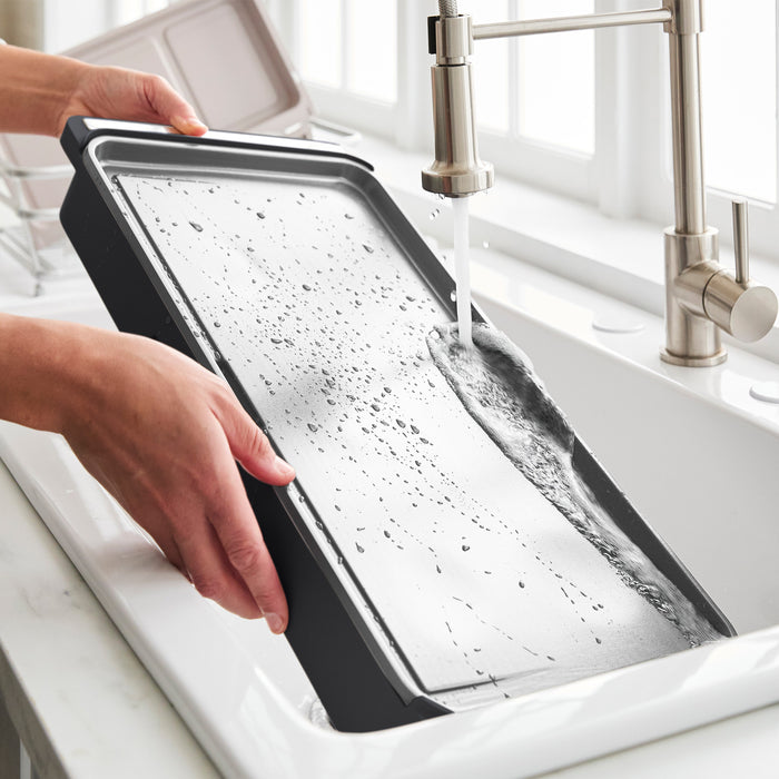 Someone washes the Advanced Griddle with Warming Drawer | Graphite under running water in a white kitchen sink after cooking a family brunch, with soap suds and droplets visible on the tray.