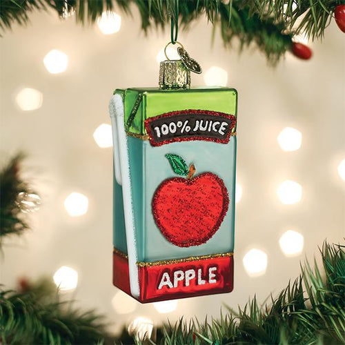 A Christmas tree ornament shaped like a juice box with a red apple illustration and the words 100% JUICE and APPLE hanging from a tree branch with blurred lights in the background.