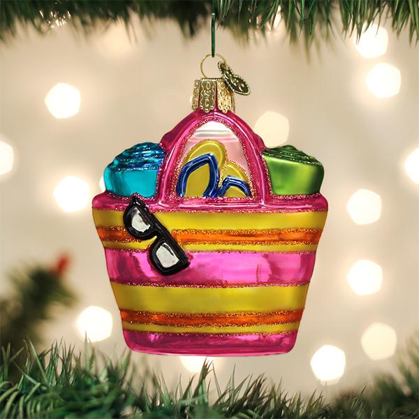 A colorful glass Christmas ornament shaped like a striped beach bag hangs on a tree, filled with flip-flops, towels, and sunglasses, with blurred Christmas lights in the background.