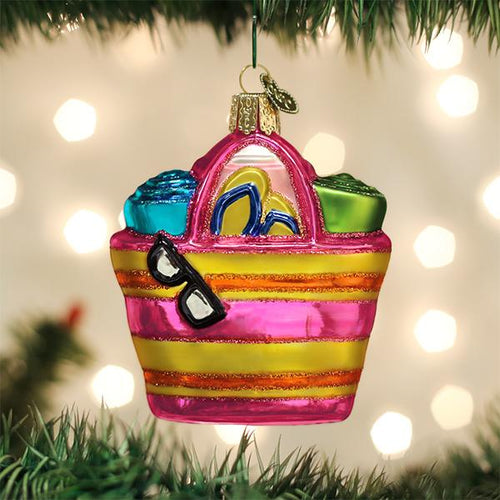 A colorful glass Christmas ornament shaped like a striped beach bag hangs on a tree, filled with flip-flops, towels, and sunglasses, with blurred Christmas lights in the background.
