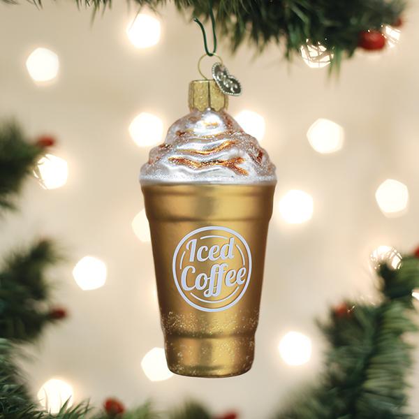 A gold, iced coffee-shaped ornament with whipped cream and glitter hangs on a Christmas tree, surrounded by green branches and blurred white lights in the background.