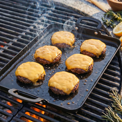 Six burger patties with melted cheese cooking on a rectangular carbon steel double burner griddle, smoke rising.