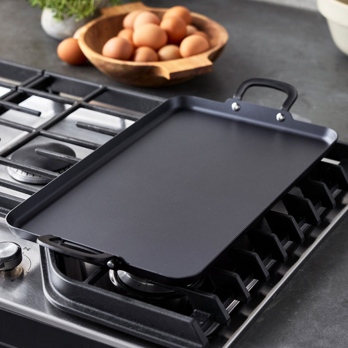 Carbon steel double burner griddle sits on a stovetop, with a wooden bowl of eggs in the background.