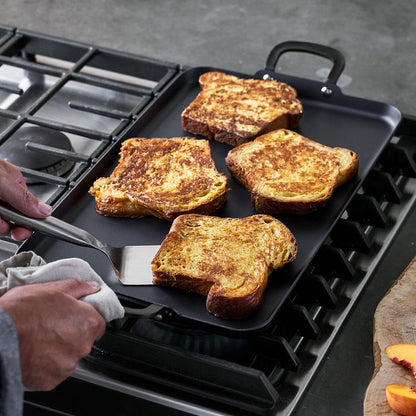 Four slices of French toast on a large black carbon steel double burner griddle, with one being flipped by a spatula.