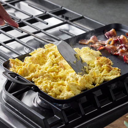 Eggs and bacon cooking on a large carbon steel double burner griddle as someone stirs the eggs.