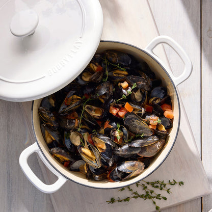 Ivory Dutch oven with mussels, tomatoes, and herbs inside, lid partially open, set on a wood surface.