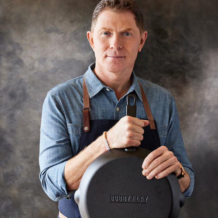 A man in a denim shirt and apron holds the Bobby Flay by GreenPan 8" and 10" Cast Iron Frypan Set, heirloom-quality skillets engraved with BOBBY FLAY, standing against a dark backdrop, ready to inspire your modern kitchen.