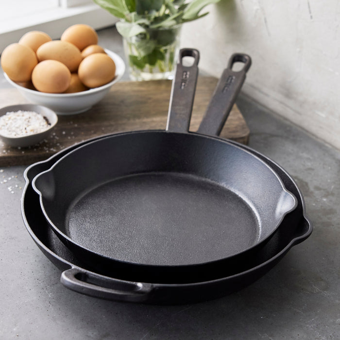 The Bobby Flay by GreenPan 8" and 10" Cast Iron Frypan Set is stacked on a gray countertop in a modern kitchen, with a bowl of eggs, salt, and fresh herbs near a window in the background.