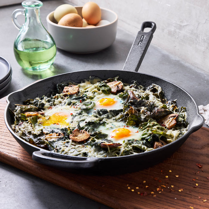 The Bobby Flay by GreenPan 8" and 10" Cast Iron Frypan Set features a spacious surface for baked eggs, sautéed greens, and mushrooms, displayed on a wooden board alongside a glass bottle with green liquid and a bowl of eggs in a modern kitchen.