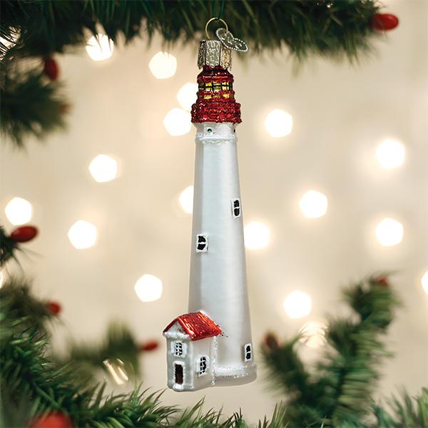 A glass Christmas tree ornament shaped like a white lighthouse with a red roof, hanging on a green tree branch with warm, blurred lights in the background.