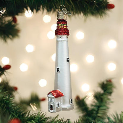 A glass Christmas tree ornament shaped like a white lighthouse with a red roof, hanging on a green tree branch with warm, blurred lights in the background.
