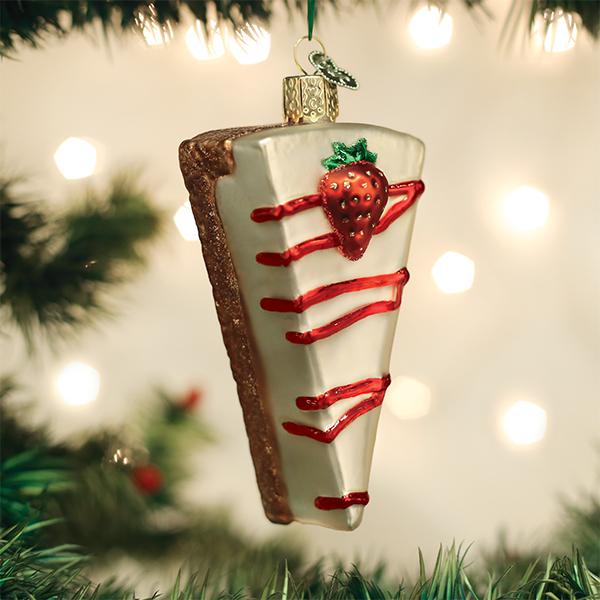 A Christmas tree ornament shaped like a slice of cheesecake with white icing, red drizzle, and a decorative strawberry, hanging among green pine branches with blurred lights in the background.