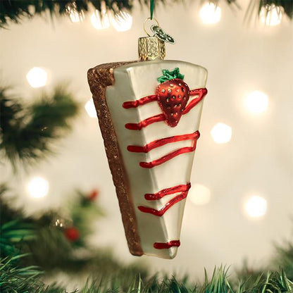A Christmas tree ornament shaped like a slice of cheesecake with white icing, red drizzle, and a decorative strawberry, hanging among green pine branches with blurred lights in the background.