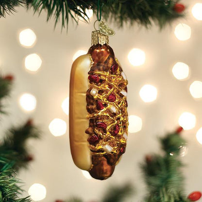 A hot dog-shaped ornament decorated with glitter and beads hangs from a Christmas tree branch, with blurred white holiday lights in the background.