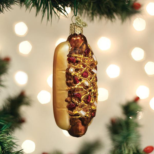A hot dog-shaped ornament decorated with glitter and beads hangs from a Christmas tree branch, with blurred white holiday lights in the background.