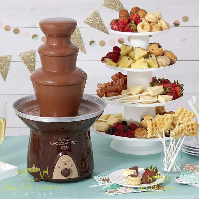 Chocolate fountain with 2 bags of chocolate rental displayed at a festive event with fruit and snacks.