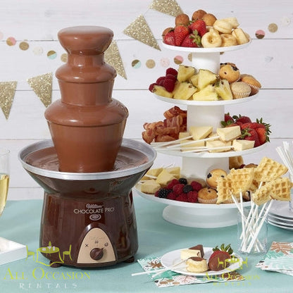 Chocolate fountain with 2 bags of chocolate rental displayed at a festive event with fruit and snacks.