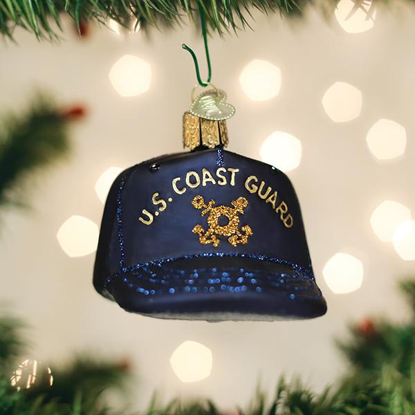 A Christmas tree ornament shaped like a navy blue baseball cap with â€œU.S. COAST GUARDâ€_x009d_ and an emblem in gold letters, hanging on a tree branch with blurred lights in the background.