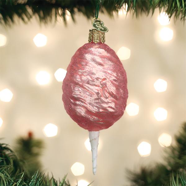 A pink, glittery cotton candy-shaped ornament hangs from a green Christmas tree branch, with soft white lights blurred in the background.