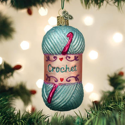 A Christmas tree ornament shaped like a blue skein of yarn with pink crochet hooks and a label that says Crochet, hanging among green branches with blurred lights in the background.