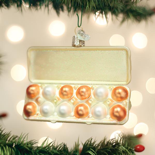 A Christmas tree ornament shaped like an open egg carton hangs on a branch, containing shiny gold and white spherical ornaments instead of eggs, with blurred lights in the background.