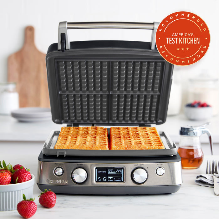 Graphite ceramic 4-square waffle maker on a counter with two cooked Belgian waffles and sliced strawberries.
