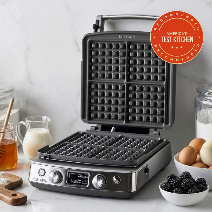 The Elite Ceramic Nonstick 4-Square Waffle Maker | Premiere Stainless Steel sits open on a kitchen counter with ingredients like eggs, milk, flour, blackberries, and honey—perfect for fluffy Belgian waffles. An Americas Test Kitchen Recommended badge is at the top right.