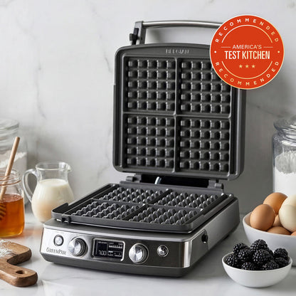 The Elite Ceramic Nonstick 4-Square Waffle Maker | Premiere Stainless Steel sits open on a kitchen counter with ingredients like eggs, milk, flour, blackberries, and honey—perfect for fluffy Belgian waffles. An Americas Test Kitchen Recommended badge is at the top right.