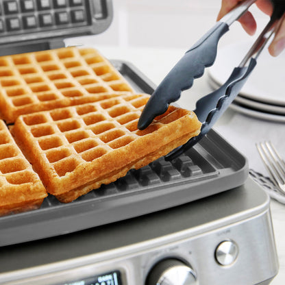 Using tongs, someone lifts a golden Belgian waffle from the Elite Ceramic Nonstick 4-Square Waffle Maker | Premiere Stainless Steel, with fresh waffles inside and plates and utensils visible in the background.