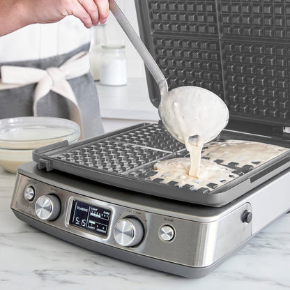 A person ladles batter onto the preheated Elite Ceramic Nonstick 4-Square Waffle Maker | Premiere Stainless Steel, featuring a digital display and control knobs, as a bowl of batter sits on the kitchen counter.