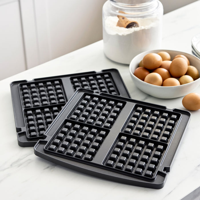 Gray multi grill and griddle with waffle plates on a countertop beside brown eggs and a jar of flour.