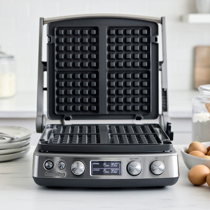 Open multi grill and griddle with waffle plates on a countertop next to eggs and flour.