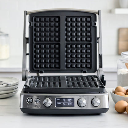 Open multi grill and griddle with waffle plates on a countertop next to eggs and flour.