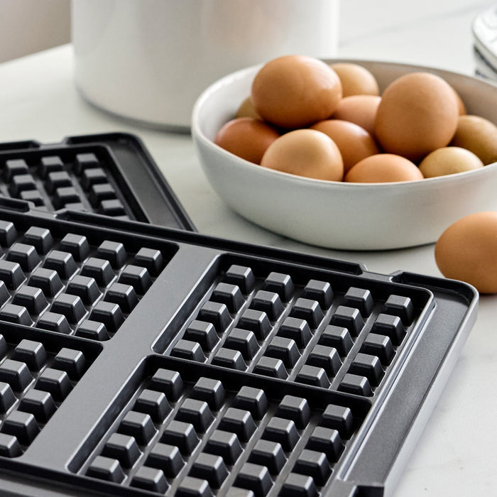 Waffle plates for a multi grill and griddle on a table next to a white bowl of brown eggs.
