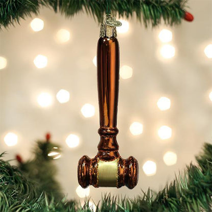 A shiny, brown gavel-shaped ornament hangs from a green Christmas tree branch with blurred white lights in the background.