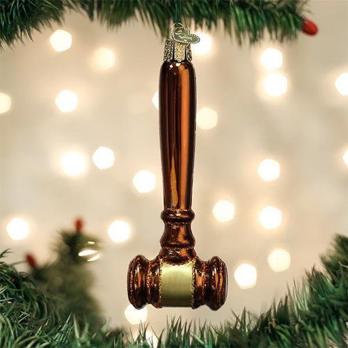 A shiny, brown gavel-shaped ornament hangs from a green Christmas tree branch with blurred white lights in the background.