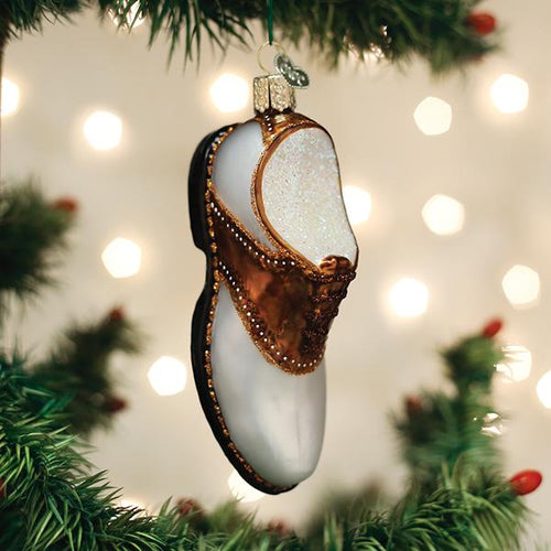 A Christmas tree ornament shaped like a traditional brown and white leather clog shoe hangs among green pine branches, with blurred festive lights in the background.