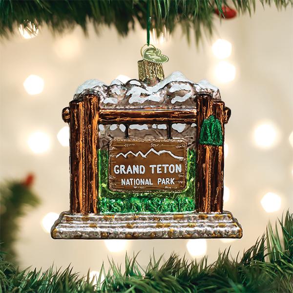 A glass ornament shaped like the Grand Teton National Park entrance sign hangs on a Christmas tree, with blurred white lights in the background.