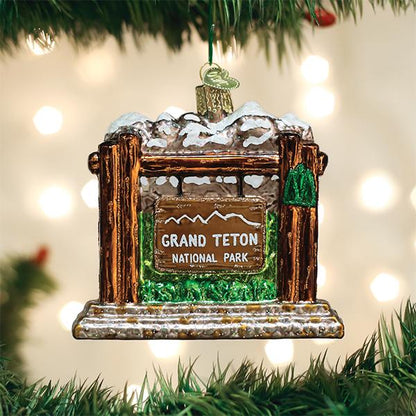 A glass ornament shaped like the Grand Teton National Park entrance sign hangs on a Christmas tree, with blurred white lights in the background.