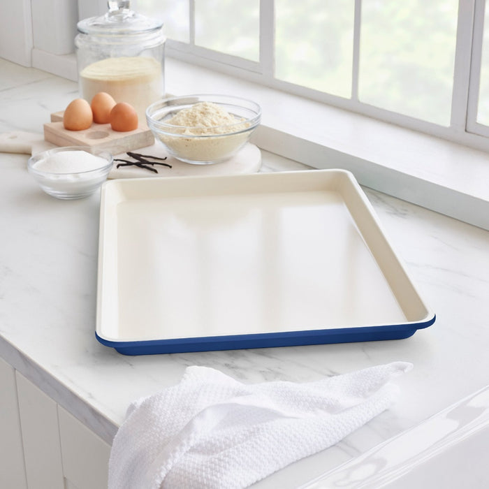 Navy ceramic nonstick cookie sheet on a kitchen counter with baking ingredients and a dish towel.