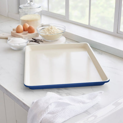 Navy ceramic nonstick cookie sheet on a kitchen counter with baking ingredients and a dish towel.